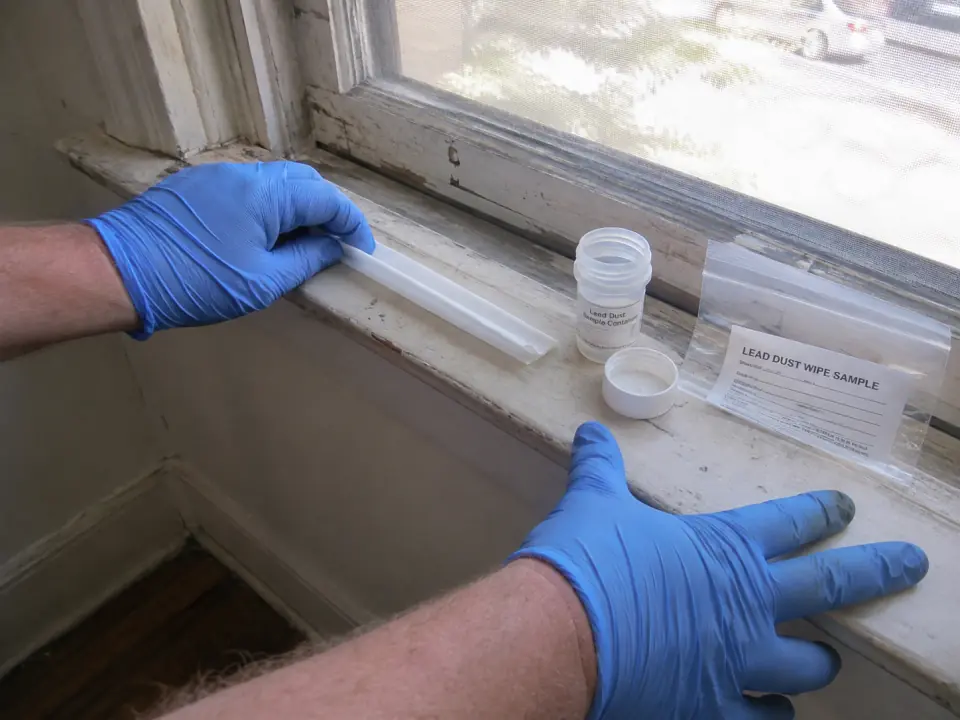 Gloved hands wiping a window sill with a lead dust sample wipe, next to an open sample container and a labelled sample bag